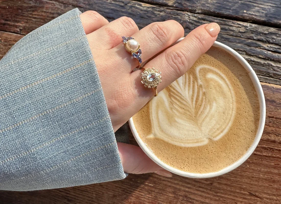 A hand wearing two rings resting on a coffee cup with latte art on a wooden surface.