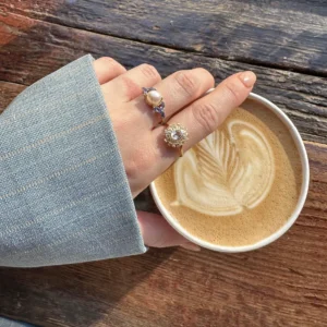 A hand wearing two rings resting on a coffee cup with latte art on a wooden surface.