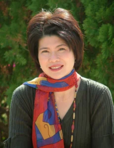 Warm portrait of Matta-Nandrakwang with short dark hair, wearing a red and blue patterned scarf and dark cardigan against lush green foliage outdoors.